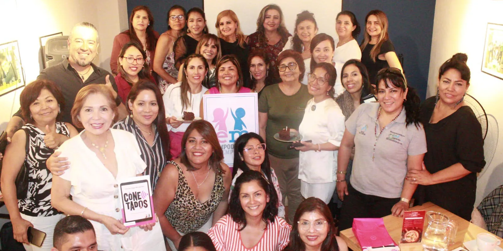 Grupo numeroso de empresarias posando en un local con el cartel de Empodérate y Emprende y el libro Conectados