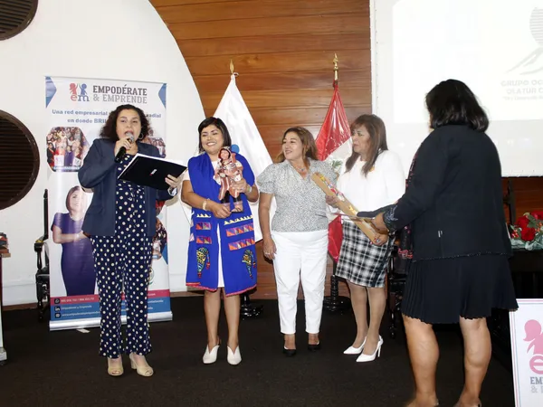 Una mujer habla al micrófono mientras otras sostienen una figura de cerámica y un obsequio en el escenario