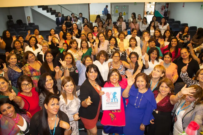 Grupo de mujeres sonriendo y saludando en una sala; al centro, un cartel de Empodérate y Emprende con Aurelia Gavidia