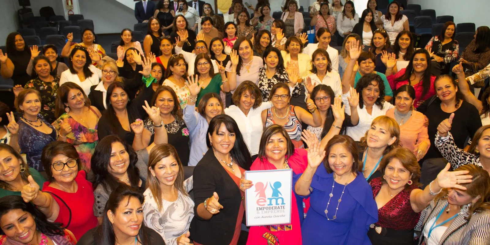 Grupo de mujeres sonriendo y saludando en una sala; al centro, un cartel de Empodérate y Emprende con Aurelia Gavidia