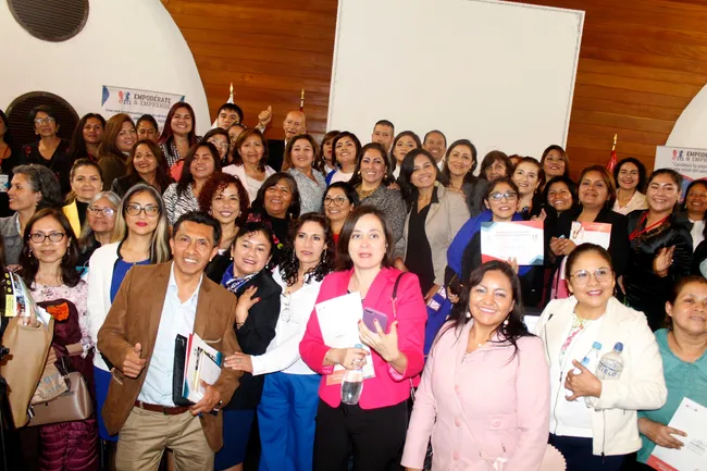 Grupo numeroso de participantes sonriendo en un salón de madera; algunas sostienen certificados y folletos