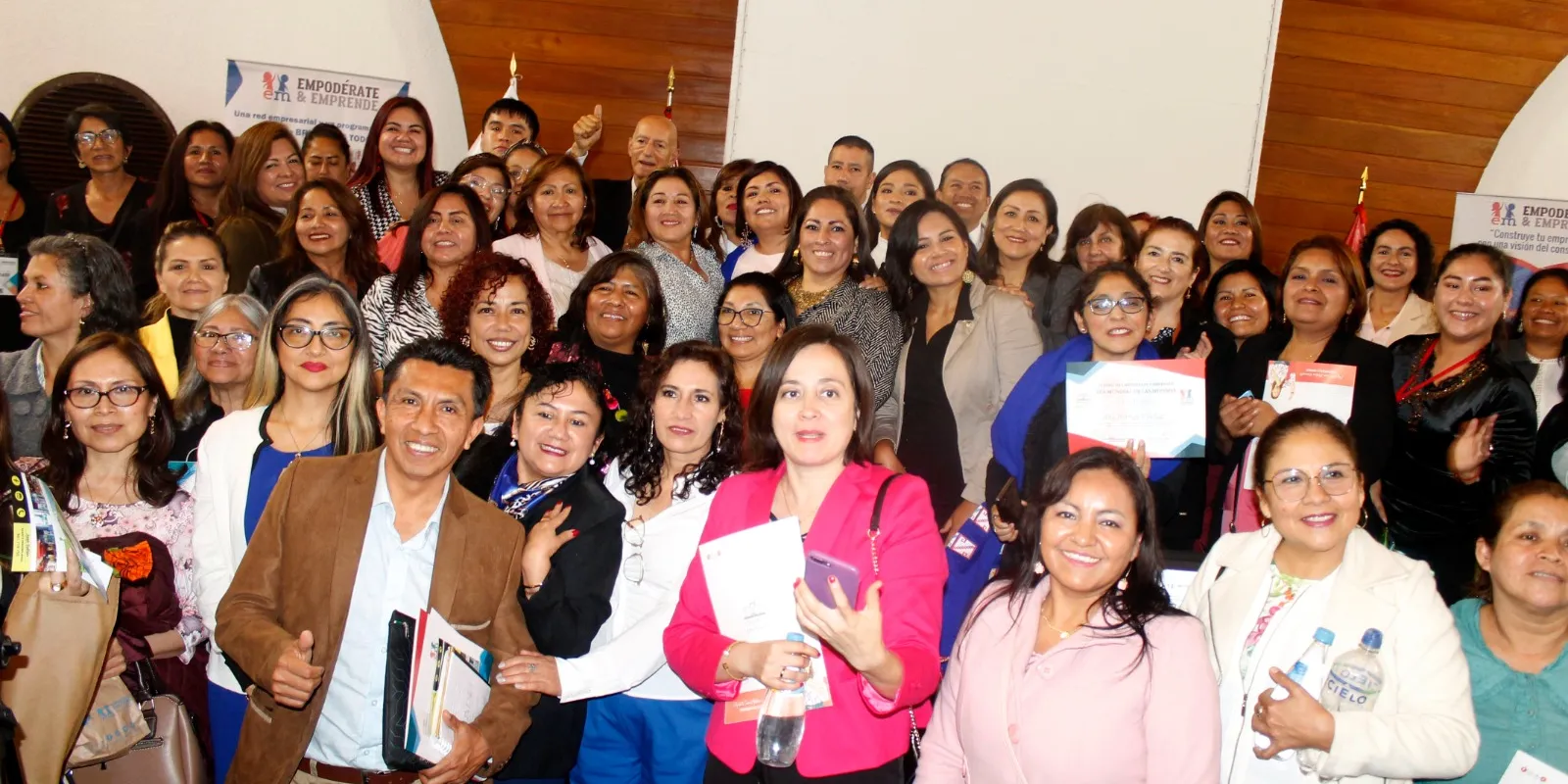 Grupo numeroso de participantes sonriendo en un salón de madera; algunas sostienen certificados y folletos